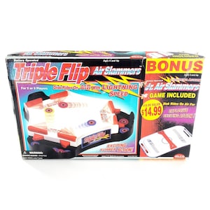 May include: A Triple Flip Air Slammers battery-operated game box featuring a white and black playing surface with red flippers. The packaging displays text including Triple Flip, Air Slammers, and Bonus Jr. Air Slammers game included.
