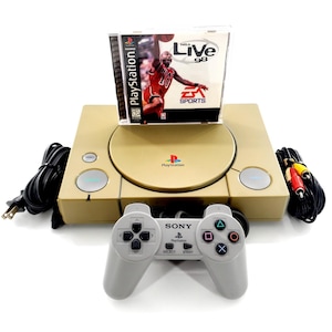 May include: A gold-colored PlayStation console with a game disc of NBA Live 98. A gray controller is in front of the console. Cables and connectors are visible on the sides. The game disc features a basketball player.