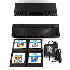 May include: An Atari 5200 video game console in black and silver with a power adapter and four game cartridges labeled Pac-Man, Defender, Joust, and Qix. The console features a rectangular design with a prominent silver horizontal stripe.