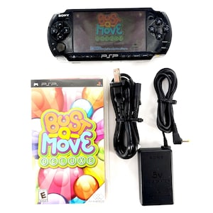 Puede incluir: Consola de juegos portátil Sony PSP negra con el juego "Bust-a-Move Deluxe" y accesorios. La caja del juego presenta burbujas coloridas. Incluye un adaptador de corriente y un cable de carga.
