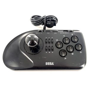 Könnte beinhalten: Ein schwarzer Sega-Arcade-Gamecontroller mit einem großen kugelförmigen Joystick auf der linken Seite, sechs Hauptaktionstasten mit der Beschriftung A, B, C, X, Y und Z, einer Start-Taste, einer Mode-Taste und einem Mega-Fire-Geschwindigkeitsregler. Ein schwarzes Kabel ist angeschlossen.