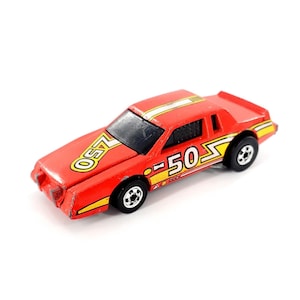 Puede incluir: Un coche de carreras de juguete rojo con detalles amarillos y blancos. El coche tiene el número "50" y la palabra "LASO" en amarillo. Tiene ruedas negras y un parabrisas negro. Es un juguete vintage.
