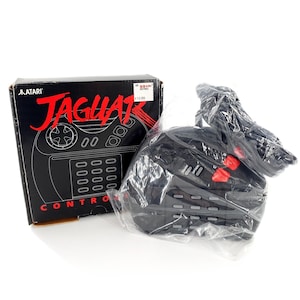 May include: A black Atari Jaguar video game controller with a numeric keypad, red action buttons, and a directional pad, partially wrapped in clear plastic, next to its original black cardboard box featuring the Atari Jaguar logo and text.