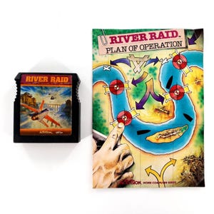 May include: A black River Raid video game cartridge for the Atari 2600 and a matching paper instruction manual titled River Raid Plan of Operation, featuring a blue river map with red target icons and a hand illustration on the cover.