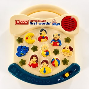 May include: A VTech Little Smart First Words Plus toy in cream, blue, and red. The toy features a handle, speaker, and a selection of colorful character buttons, including a moon, bird, and dog. The toy has a curved base with musical notes.
