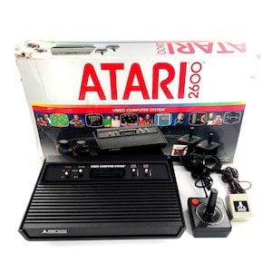 May include: An Atari 2600 video game console set, including the console, joysticks, and game cartridges. The console is black with a ribbed design and the Atari logo. The box is white with red "ATARI 2600" text.