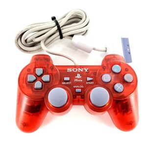 Puede incluir: Un mando de juego Sony PS one rojo translúcido con joysticks analógicos grises, una cruceta y botones de acción. El mando incluye un cable gris conectado con un enchufe blanco y una brida negra que sujeta el cable.