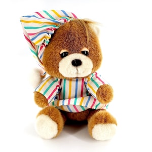 Puede incluir: Un oso de peluche marrón con gorro de dormir a rayas y pijama a juego. El oso tiene hocico y patas blancos. El pijama y el gorro presentan rayas verticales de colores rosa, azul, amarillo y verde. El oso está sentado.
