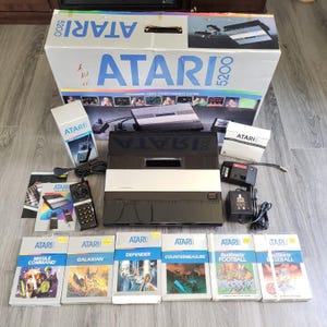 May include: An Atari 5200 video game console set including the main black and silver console, a controller, power supply, RF switch, and six game boxes: Missile Command, Galaxian, Defender, Countermeasure, RealSports Football, and Baseball.