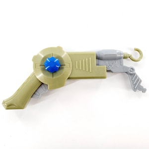 May include: A tan and gray toy gun with a blue center. The toy gun has a hook on the end and the text "BIG GUY" on the side. The toy gun is made of plastic.
