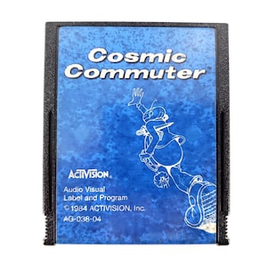May include: A black rectangular video game cartridge with a blue label featuring the text Cosmic Commuter, an illustration of a character in motion, the Activision logo, and the text Audio Visual Label and Program 1984 ACTIVISION, Inc. AG-038-04.
