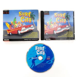 May include: A Philips CD-i Kids Surf City interactive game set including a cardboard outer sleeve, a plastic jewel case, and a blue compact disc. The cover art features a red convertible car with cartoon characters, a palm tree, and the text Surf City.