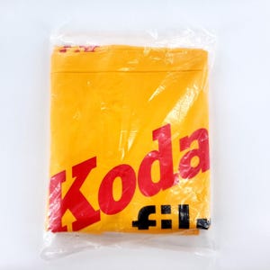 May include: A folded yellow textile item encased in clear plastic packaging. The fabric features the red text 'Koda' in a bold, sans-serif font, with the black text 'film' positioned directly below it in a smaller, lowercase typeface.