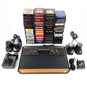 Könnte beinhalten: Eine schwarze Atari Video Computer System Konsole mit Holzmaserung, begleitet von drei schwarzen Joysticks, zwei Paddle-Controllern, einem Netzteil und einer großen Sammlung gestapelter rechteckiger Spielkassetten mit verschiedenen Titeln.