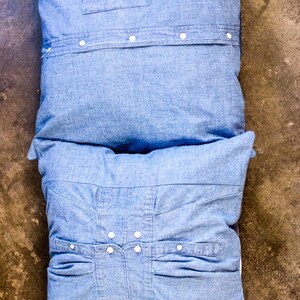 May include: Two blue denim pillows with button details. The top pillow has a single pocket and the bottom pillow has two pockets.