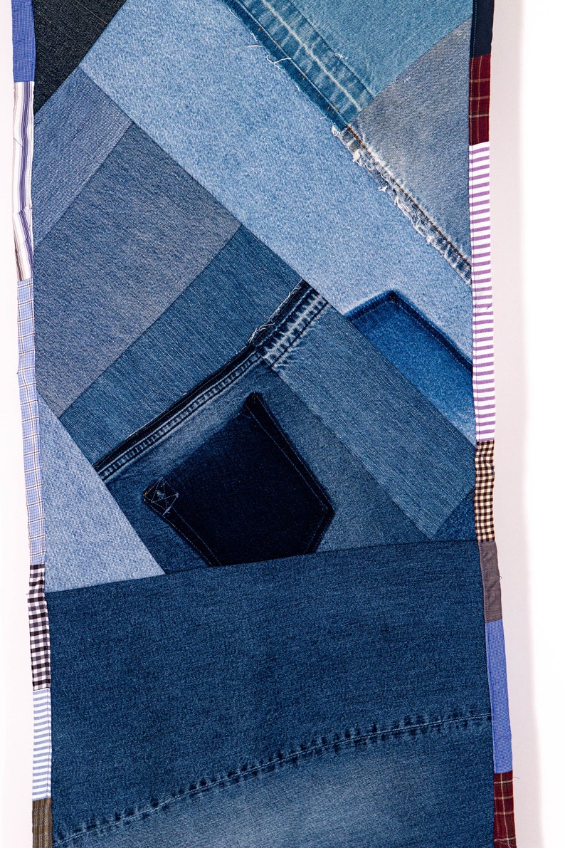May include: A patchwork quilt made from different shades of blue denim fabric. The quilt is made up of various sized squares and rectangles, with a pocket sewn into one of the squares. The quilt is edged with a variety of patterned fabrics, including plaid, striped, and solid colors.