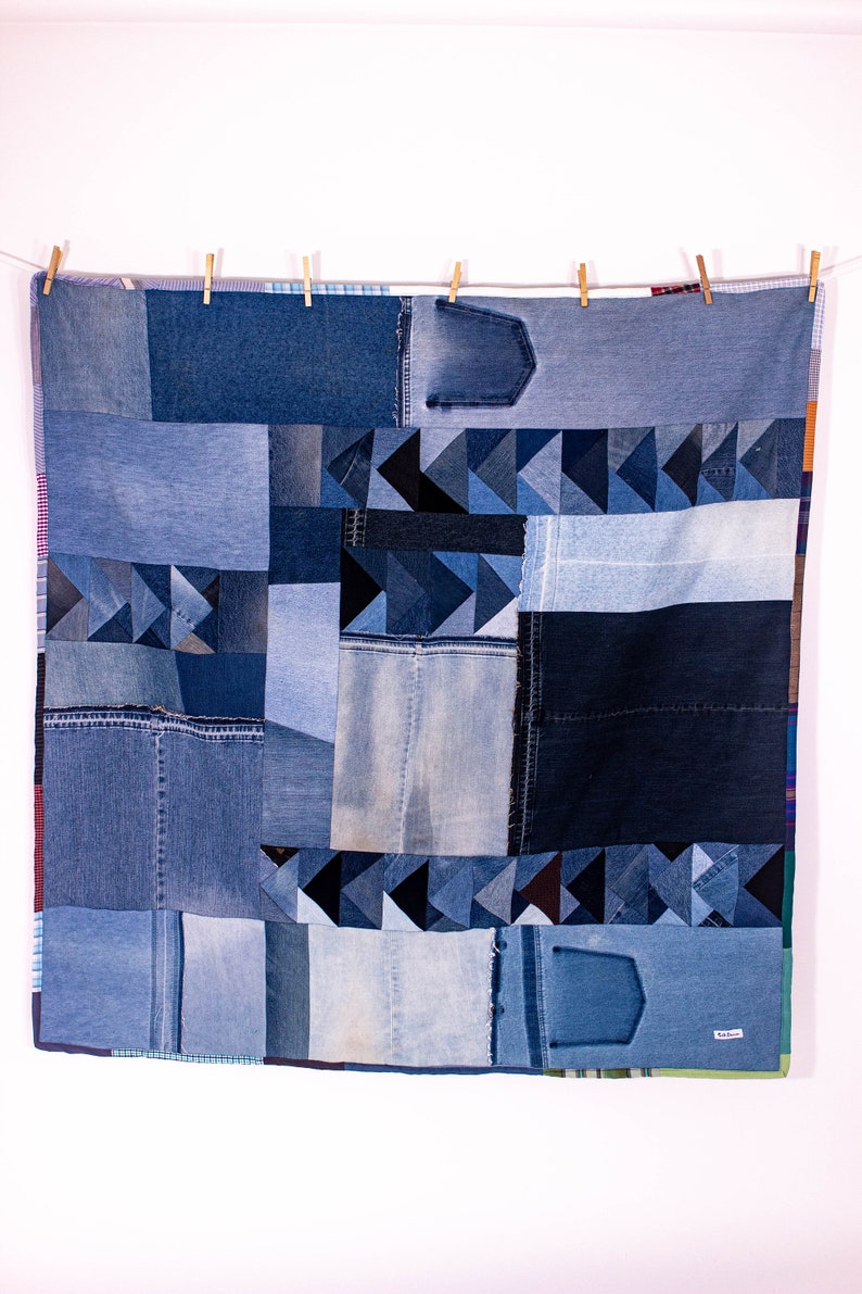 May include: A blue denim quilt made from repurposed jeans. The quilt is made up of various sized squares and triangles of denim fabric. The quilt is hanging on a clothesline with wooden clothespins.