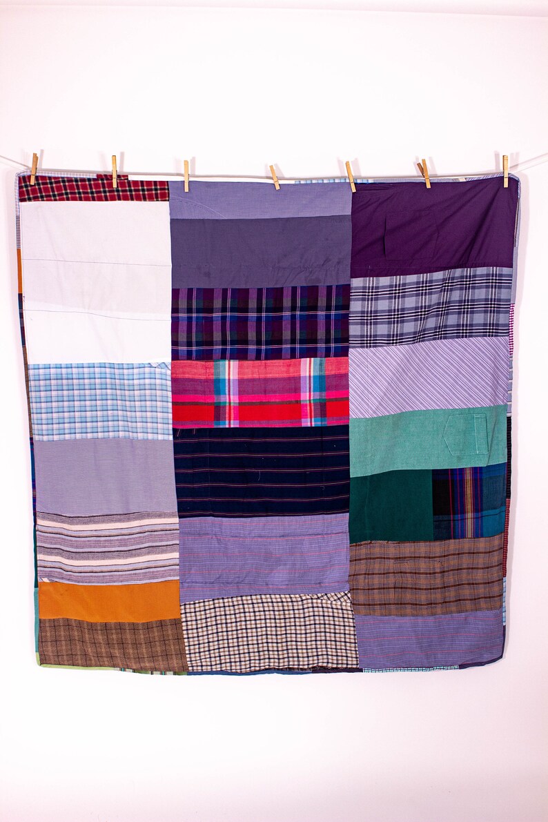 May include: A patchwork quilt made from repurposed fabric scraps. The quilt is made up of squares of different colors and patterns, including plaid, striped, and solid colors. The quilt is hanging on a clothesline.