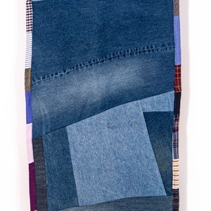 May include: A patchwork quilt made from different shades of blue denim fabric. The quilt is made up of various sized squares and rectangles, creating a geometric pattern. The edges of the quilt are trimmed with a pink, purple, and brown plaid fabric.