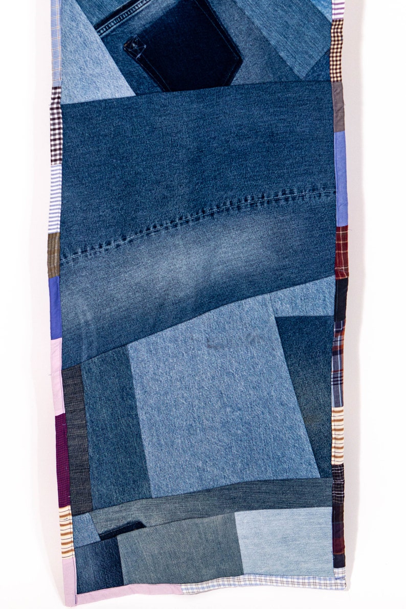 May include: A patchwork quilt made from different shades of blue denim fabric. The quilt is made up of various sized squares and rectangles, creating a geometric pattern. The edges of the quilt are trimmed with a pink, purple, and brown plaid fabric.