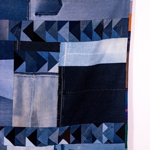 May include: A patchwork quilt made from different shades of blue denim. The quilt is hanging on a clothesline with wooden clothespins. The quilt is made up of squares and triangles of denim, and some of the squares have pockets. The quilt has a label that says "Ed Dunn" in the bottom right corner.