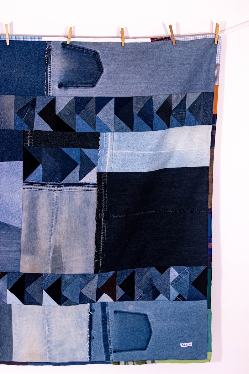 May include: A patchwork quilt made from different shades of blue denim. The quilt is hanging on a clothesline with wooden clothespins. The quilt is made up of squares and triangles of denim, and some of the squares have pockets. The quilt has a label that says "Ed Dunn" in the bottom right corner.
