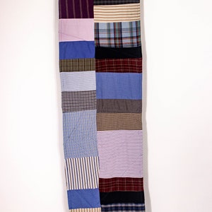 May include: A long, rectangular patchwork quilt made from various fabrics in shades of blue, brown, purple, and white. The quilt is made up of squares and rectangles of different sizes and patterns, creating a visually interesting and textured design.