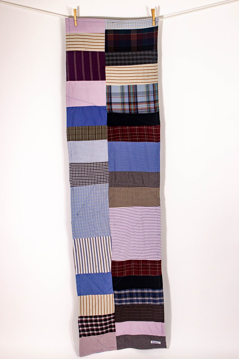 May include: A long, rectangular patchwork quilt made from various fabrics in shades of blue, brown, purple, and white. The quilt is made up of squares and rectangles of different sizes and patterns, creating a visually interesting and textured design.