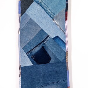 May include: A long, narrow wall hanging made from various shades of blue denim fabric. The fabric is sewn together in a patchwork pattern, creating a geometric design. The edges of the hanging are trimmed with different colored fabrics, including pink, white, and brown.