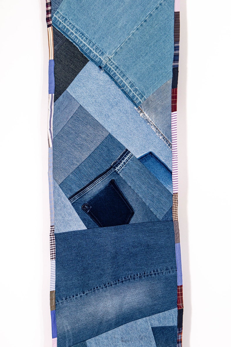 May include: A long, narrow wall hanging made from various shades of blue denim fabric. The fabric is sewn together in a patchwork pattern, creating a geometric design. The edges of the hanging are trimmed with different colored fabrics, including pink, white, and brown.