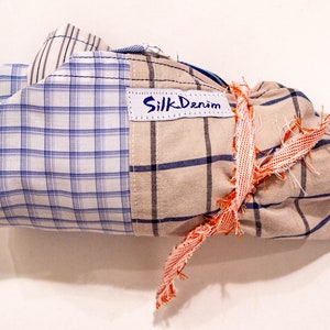 May include: A bundle of plaid fabric wrapped with a frayed orange ribbon and tied with a bow. The fabric has a blue and white plaid pattern on one side and a tan and blue plaid pattern on the other side. A white label with the text "Silk Denim" is attached to the fabric.