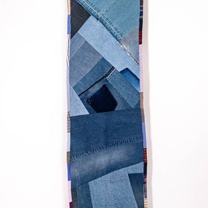 Op de afbeelding: Een lange, rechthoekige denim quilt gemaakt van verschillende tinten blauwe denim stof. De quilt is gemaakt van verschillende vierkanten en rechthoeken van denim, die aan elkaar zijn genaaid om een patchwork ontwerp te creëren. De quilt hangt aan twee houten wasknijpers aan een witte witte muur.