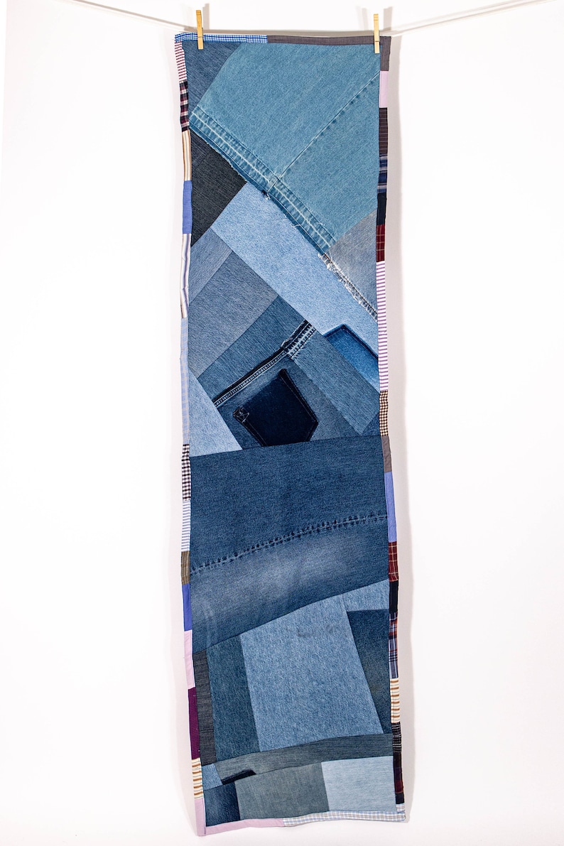 May include: A long, rectangular denim quilt made from various shades of blue denim fabric. The quilt is made up of different sized squares and rectangles of denim, sewn together to create a patchwork design. The quilt is hanging from two wooden clothespins on a white wall.