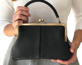 May include: A black handbag with a textured surface and a single top handle. The bag features a rectangular brass-toned metal frame with a kiss-lock closure mechanism at the top center. The design includes a vertical seam down the front.