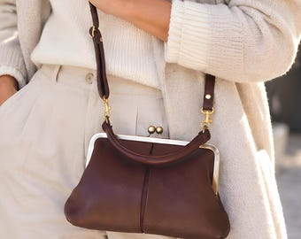 May include: A dark brown leather handbag with a silver-toned metal frame and a kiss-lock closure. The bag features a short, rounded handle and a detachable, adjustable shoulder strap with gold-toned hardware clips.