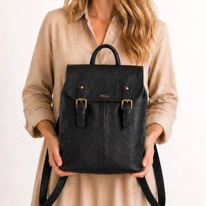 May include: Black leather backpack with a top handle and adjustable shoulder straps. The backpack features a flap closure with two buckle straps and gold-colored hardware. The initials "TKA" are embossed on the front.