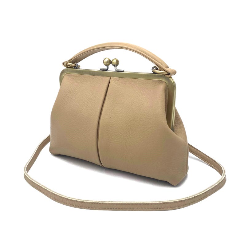 May include: A beige leather handbag featuring a structured frame with a brass-toned metal kiss-lock closure. The bag includes a single top handle and a thin, detachable shoulder strap, showcasing a soft, pebbled texture and a central vertical seam.