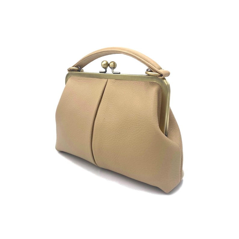 May include: A beige leather handbag with a textured surface, featuring a structured brass-toned metal frame, a top handle, and a dual-ball clasp closure. The bag has a soft, rounded silhouette with a central vertical seam on the front panel.