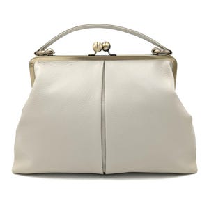 Puede incluir: Un bolso de mano de cuero blanco con superficie texturizada, que presenta una estructura r&iacute;gida, un asa superior &uacute;nica y un cierre met&aacute;lico de boquilla. El bolso tiene una costura vertical central y una silueta suave y redondeada.