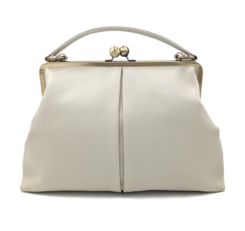 Puede incluir: Un bolso de mano de cuero blanco con superficie texturizada, que presenta una estructura r&iacute;gida, un asa superior &uacute;nica y un cierre met&aacute;lico de boquilla. El bolso tiene una costura vertical central y una silueta suave y redondeada.