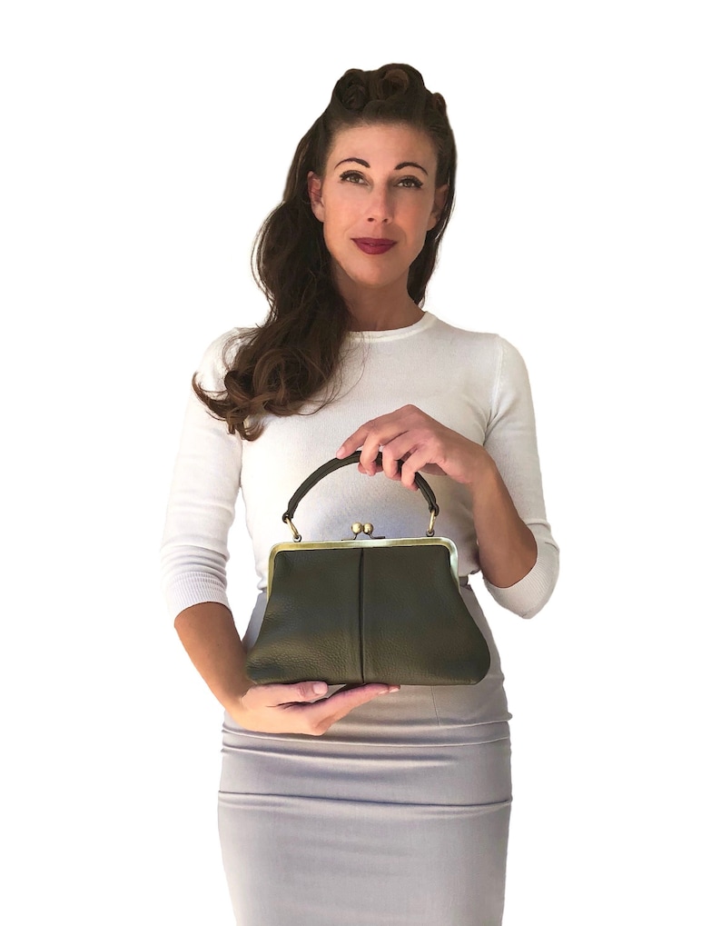 May include: A woman holds a dark green leather handbag with a gold clasp and a top handle. The bag has a slightly rounded shape and a subtle texture.