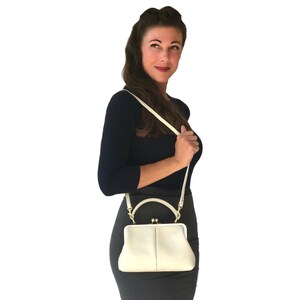 May include: A cream-colored handbag with a structured frame, a top handle, and a detachable shoulder strap. The bag features a kiss-lock closure mechanism with gold-toned metal hardware, a smooth exterior finish, and a rectangular base shape.