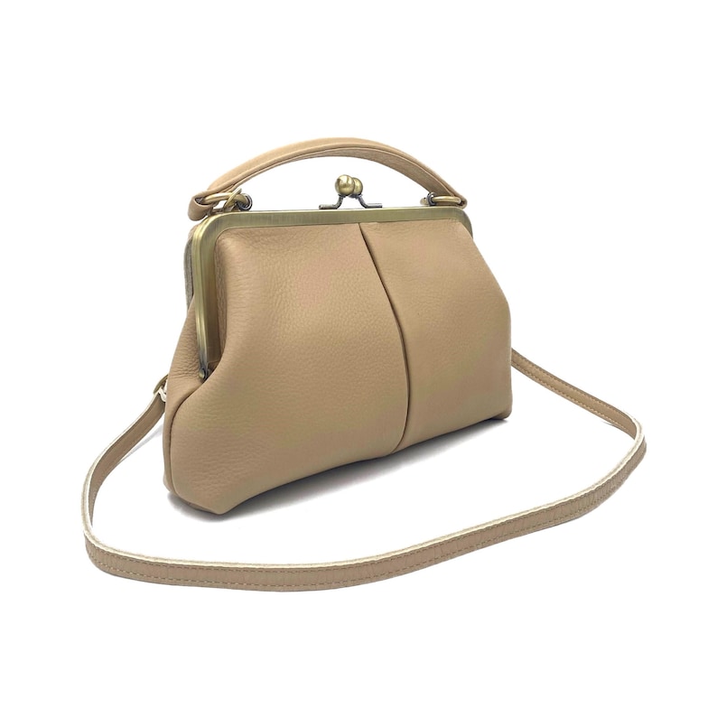 May include: A beige leather handbag with a textured surface, featuring a structured brass-toned metal frame, a top handle, and a thin matching shoulder strap. The bag has a kiss-lock closure mechanism at the top center and a soft, rounded shape.