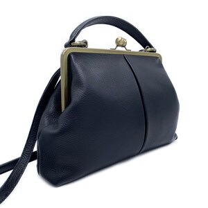 Puede incluir: Un bolso de mano azul marino oscuro con superficie texturizada, que presenta un cierre de marco de metal en tono lat&oacute;n con un broche central de bola. El bolso incluye un asa superior &uacute;nica y una correa de hombro delgada de dise&ntilde;o limpio.