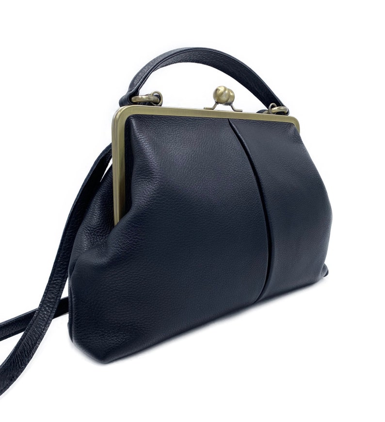 Puede incluir: Un bolso de mano azul marino oscuro con superficie texturizada, que presenta un cierre de marco de metal en tono lat&oacute;n con un broche central de bola. El bolso incluye un asa superior &uacute;nica y una correa de hombro delgada de dise&ntilde;o limpio.