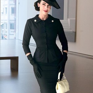 May include: A black long-sleeved jacket with a collar and small star-shaped pins, paired with a matching black pencil skirt. The outfit includes a wide-brimmed black hat, black gloves, and a small cream-colored handbag with a gold-toned clasp.