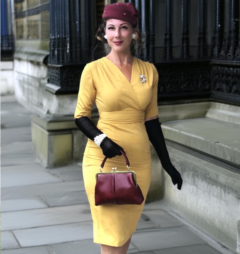 Könnte beinhalten: Eine Frau in einem gelben Kleid mit einem burgunderfarbenen Hut und schwarzen Handschuhen, die eine burgunderfarbene Handtasche hält.