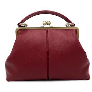 Puede incluir: Un bolso de mano de color rojo intenso con superficie texturizada, que cuenta con un marco de metal en tono lat&oacute;n y un asa superior. El bolso se asegura con un cierre de doble bola en el centro y presenta una costura vertical en el frente.