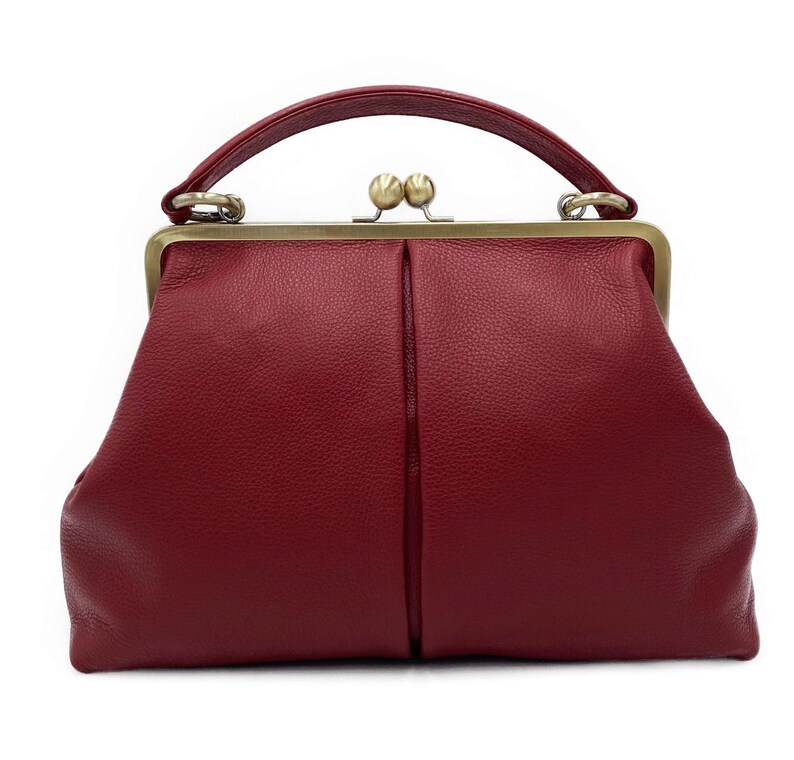 Puede incluir: Un bolso de mano de color rojo intenso con superficie texturizada, que cuenta con un marco de metal en tono lat&oacute;n y un asa superior. El bolso se asegura con un cierre de doble bola en el centro y presenta una costura vertical en el frente.