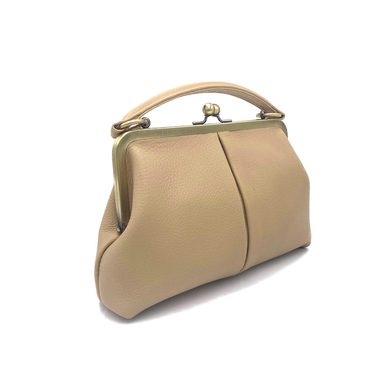 May include: A beige leather handbag with a textured surface, featuring a curved top handle and a brass-toned metal frame closure. The bag has a structured, rounded shape with a central vertical seam and a classic kiss-lock clasp mechanism.